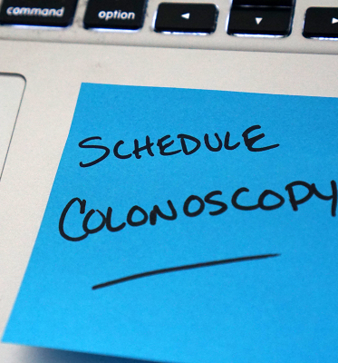 A posit note stuck to a laptop. It reads, Scheudle Colonoscopy